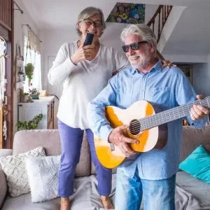 An older woman sings, and an older man plays guitar in their living room.