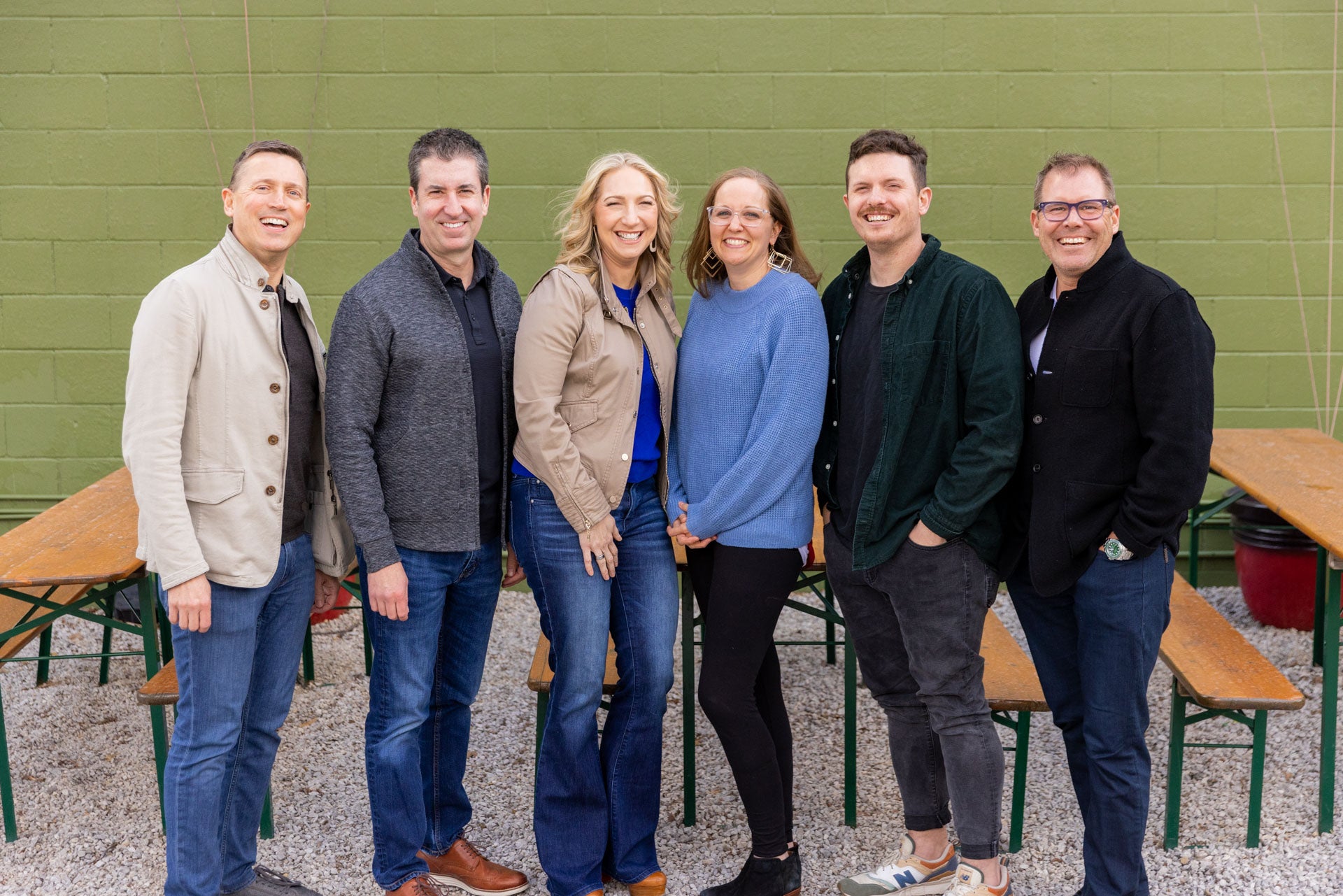 Group of six people standing together in front of a green wall outside.