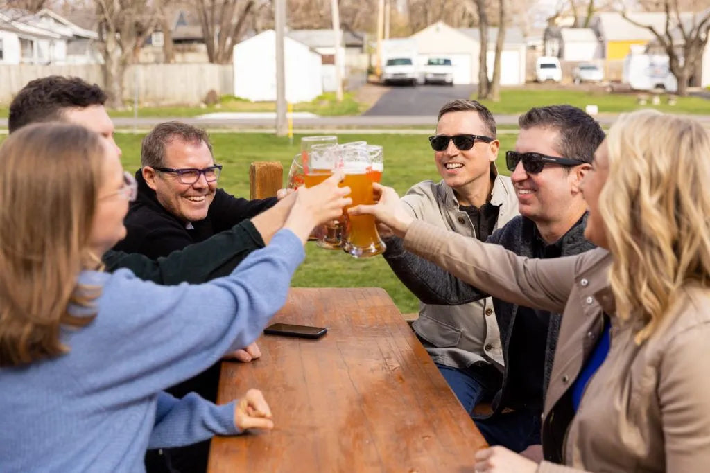Group of people toasting with beer glasses outdoors at a wooden table.