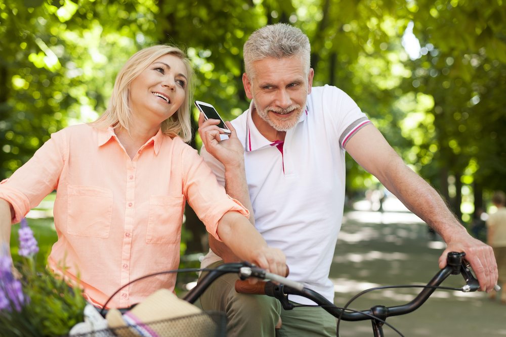 Mature couple using mobile phone while riding on bikes in park with almost invisible hearing aids