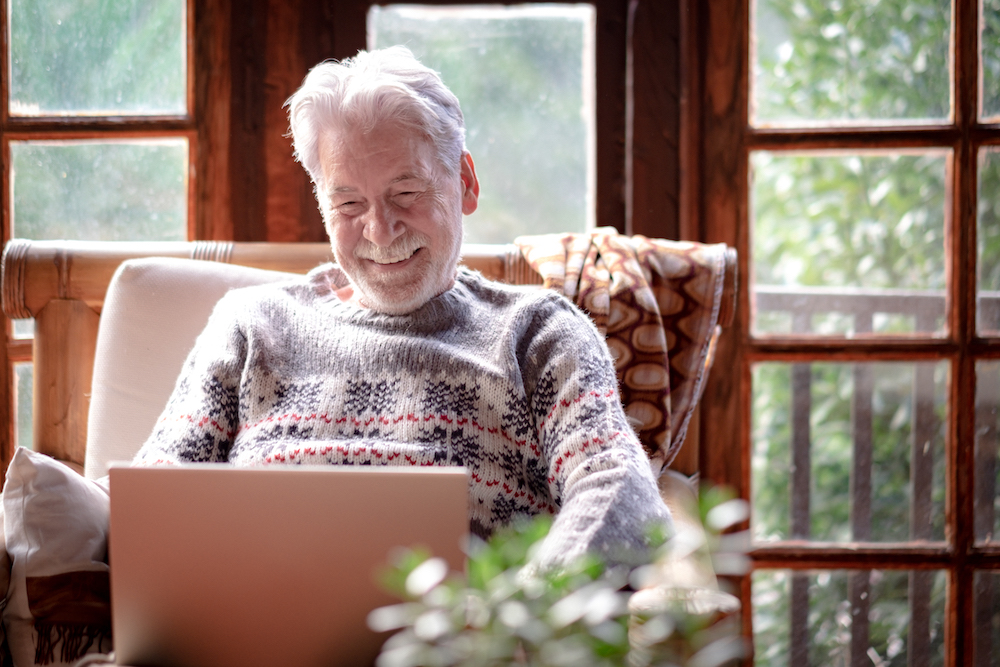 Smiling senior man in winter sweater sitting in living room using laptop computer for how do i get a hearing aid
