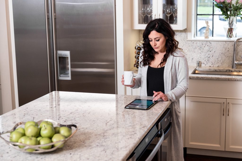 A woman drinks coffee in her kitchen while listening to her tablet. 