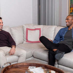 Two men laugh as they sit on a comfy couch in a well-lit room.
