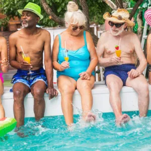 5 older people sit on the edge of a pool laughing and talking.