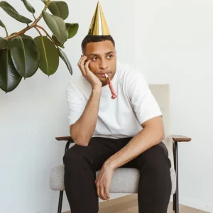 A man in a party hat sits alone in a corner looking depressed. 