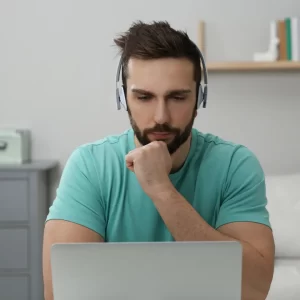 A man wearing headphones looks thoughtfully at his laptop.