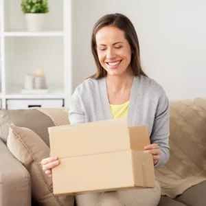 A woman smiles as she opens the cardboard box on her lap.