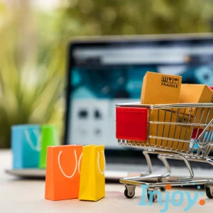 A mini shopping cart full of bags stands in front of a laptop. 