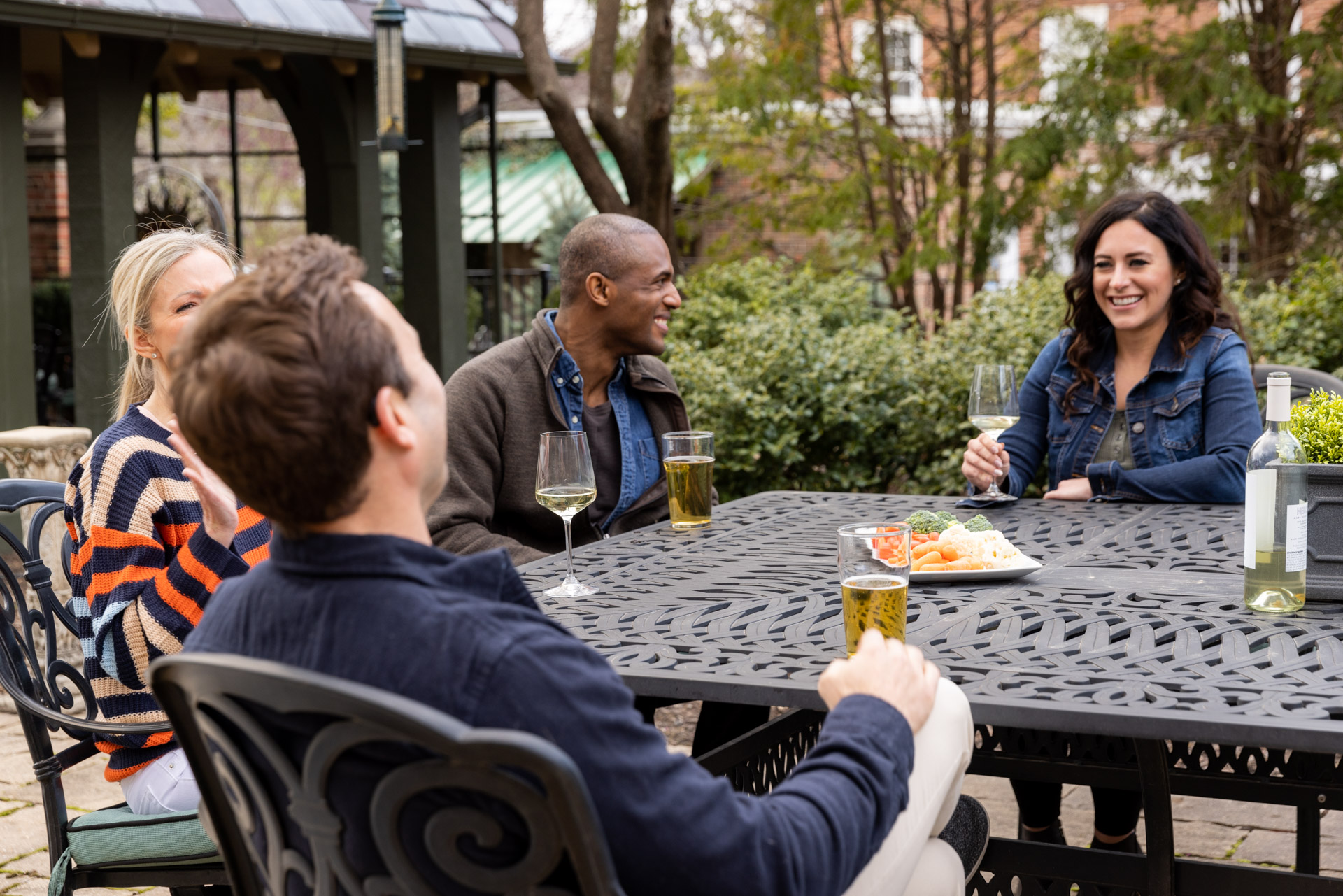 Two men and two women sitting around a table outside, talking. The men are drinking beer, and the women are drinking white wine. They are smiling. 
