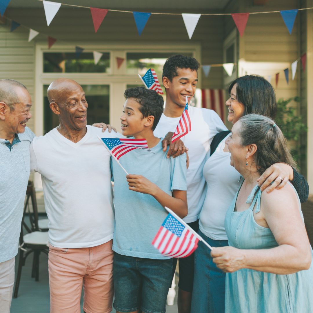 A family with two men, two teen boys, and two women stands on a front porch holding flags. They are smiling with their arms around each other’s shoulders.