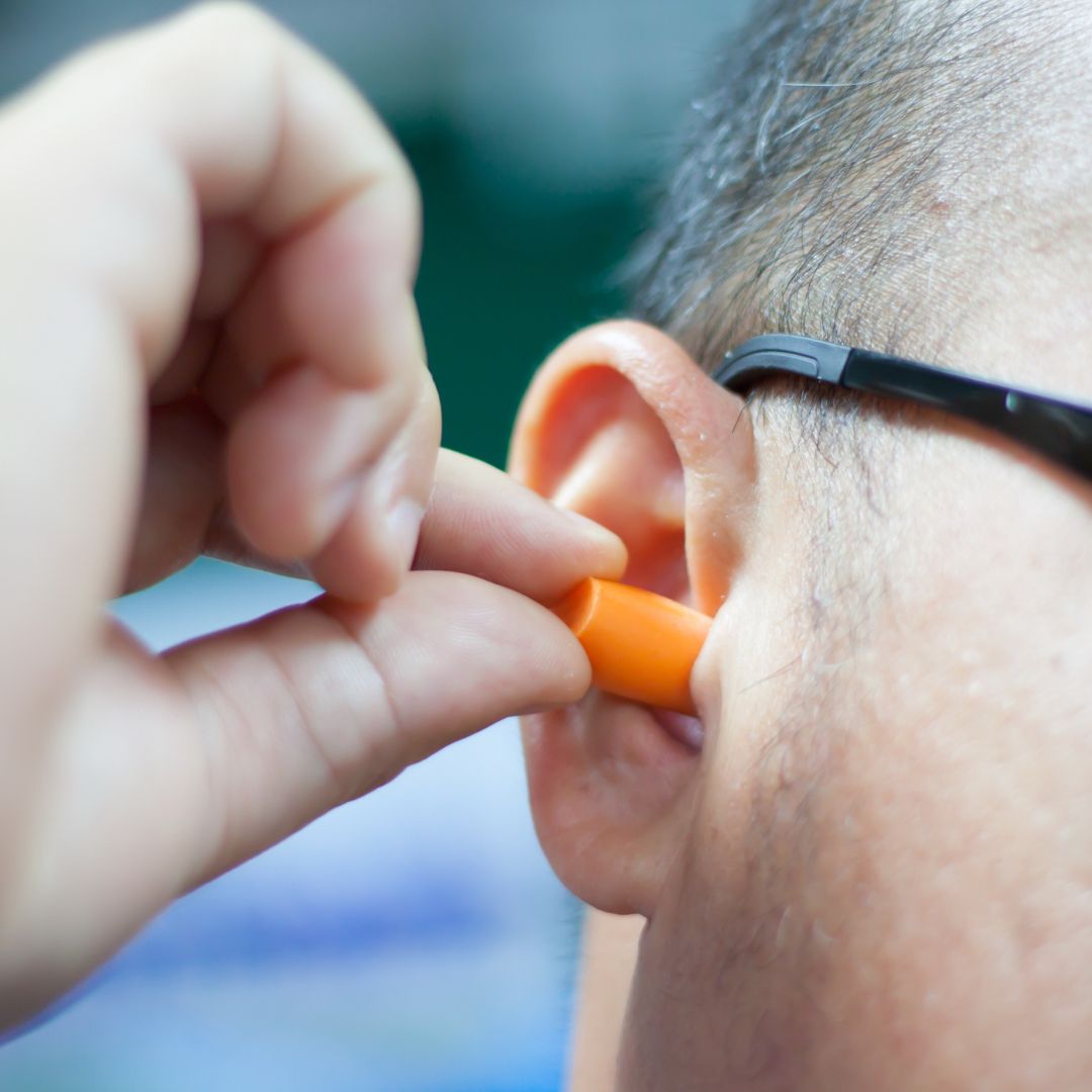 A man uses one hand to place an orange earplug into his ear. We see a bit of his face, dark hair, and glasses.