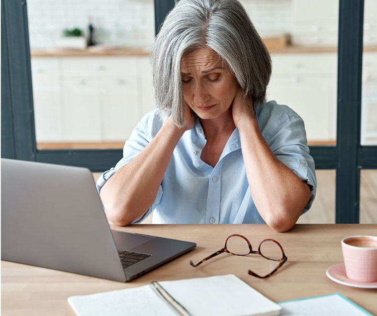 Sad older woman sits in front of her laptop and massages her neck.
