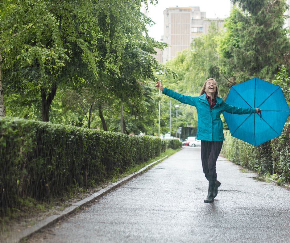 A smiling woman stands in the rain. She holds her umbrella to the side as she stretches her arms out.