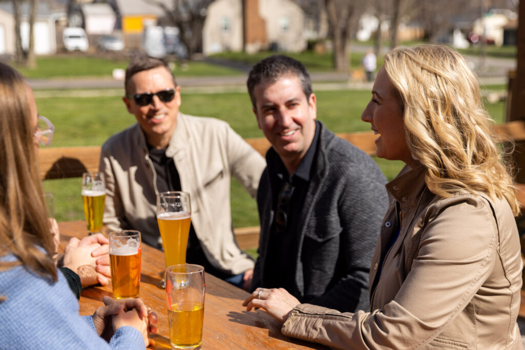 Two men face the camera, and two women are facing away. They are sitting at a table outside, talking and drinking beer.