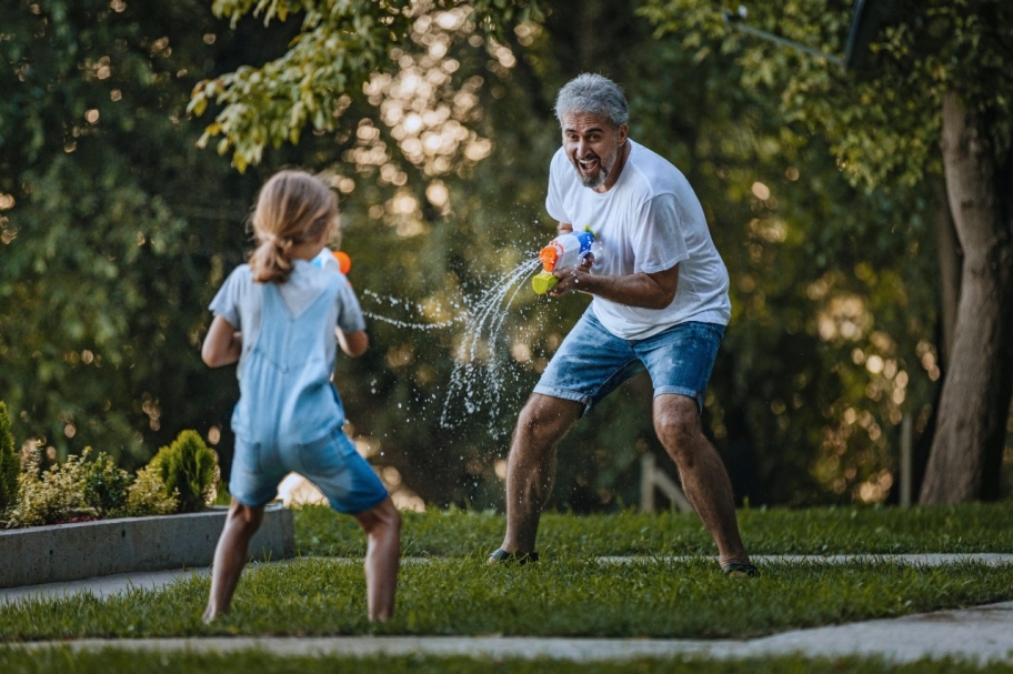 A gray-haired man laughs as he has a water-soaker fight with a child on a sunny day.