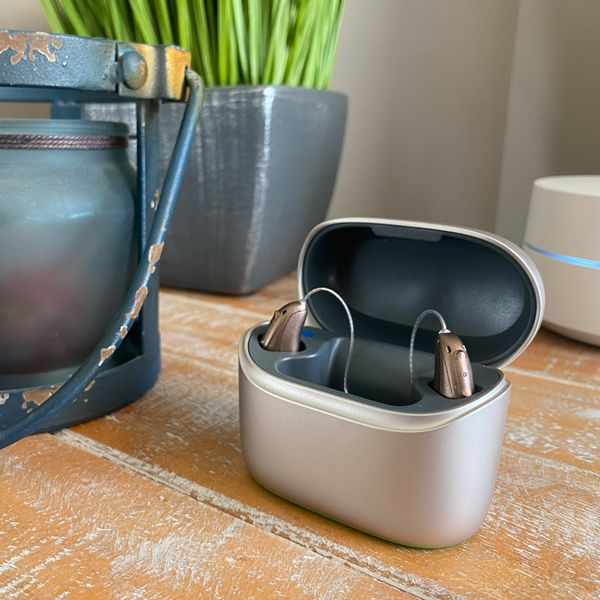 Metallic gold-colored Phonak hearing aids in their open charging case sit on a table. Behind them are a speaker and a plant.