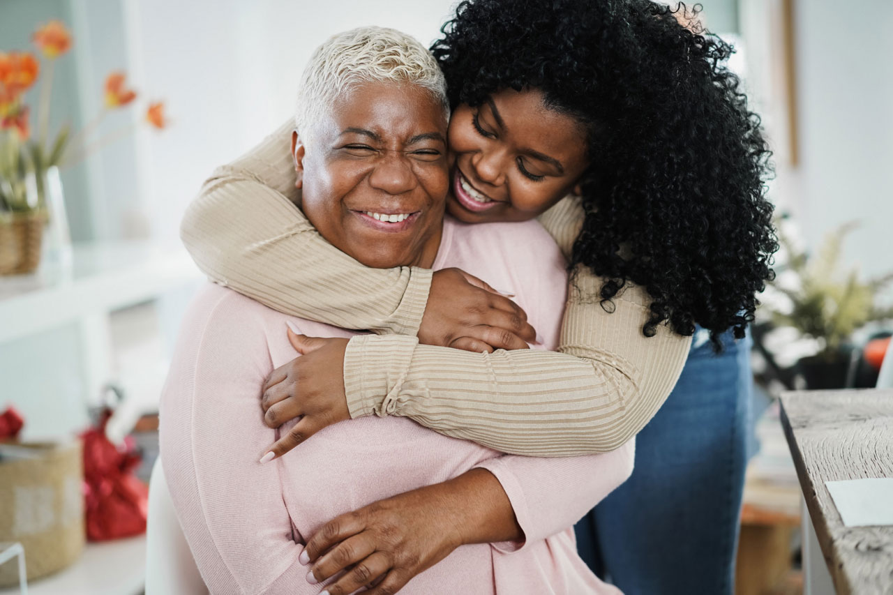 A young woman stands behind her seated mother and hugs her as both smile.