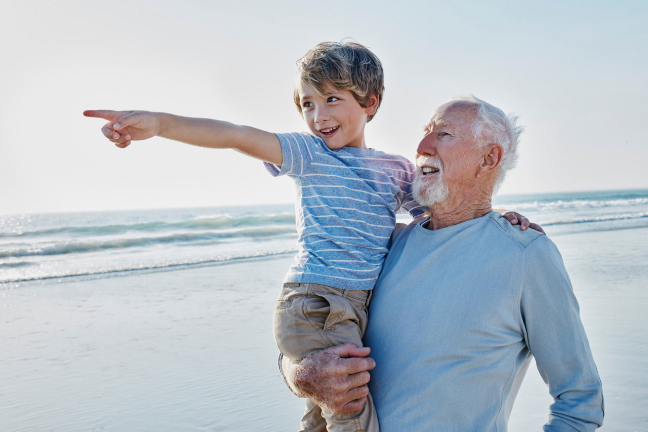 A grandfather stands and holds his happy grandson in his arm on the beach as waves roll in the background.