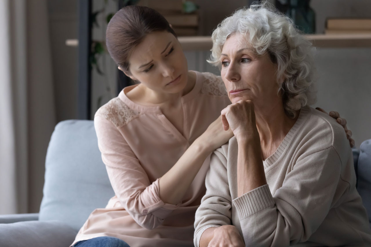 A sad, gray-haired woman sits on a couch while a younger woman tries to comfort her.