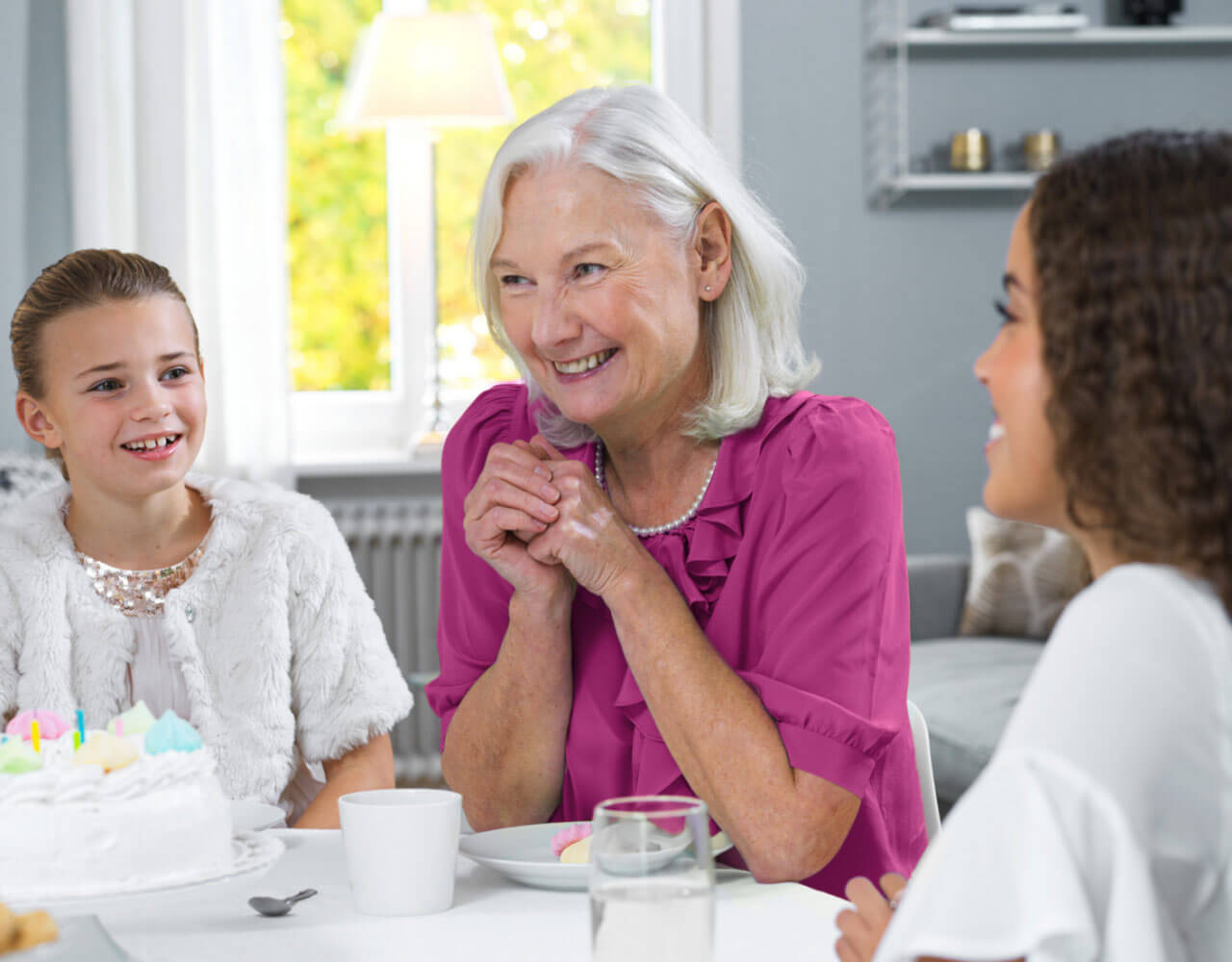 A grandmother smiles at a dining table. She sits between her two granddaughters. 