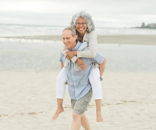 An older man carries an older woman piggyback on a beach.